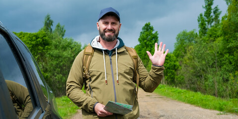Smiling friendly bearded man with backpack stands near crossover vehicle on rural road and waves hand while holding paper map. Concept of road trip, outdoor recreation and tourism. © Dmitrii Masquerade