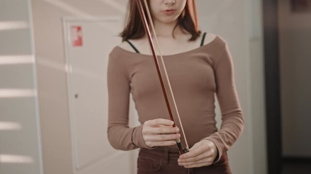 White student inspecting violin bow before practice, tightening screw, checking horsehair, applying rosin, careful hands, case open on floor, calm concentration, preparation for rehearsal