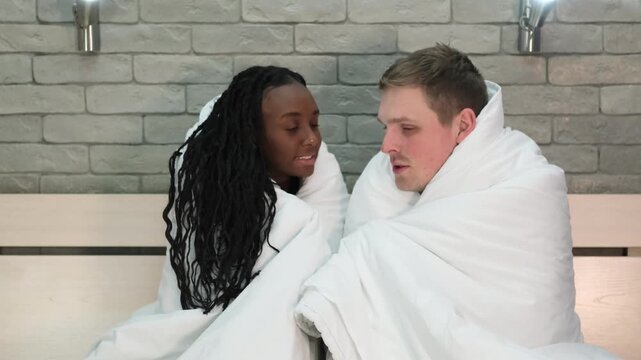 Young multiethnic couple under blanket in bed at home. People look upset and cold. A married couple is trying to stay warm at home in winter. Problems with heating your home during the cold season