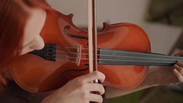 Closeup violin practice redhead student bowing in cozy music room, focused hands on fingerboard and chinrest, wooden instrument warmed by sunlight, careful bow stroke guided by teacher offcamera,