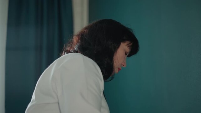 White female doctor home visit performing bedside examination with stethoscope reviewing chart and records teal curtain and window light calm professional demeanor documenting vitals advising patient