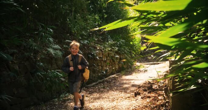 Happy boy with backpack walking down dirt path in Japanese forest with strong breeze - handheld shot