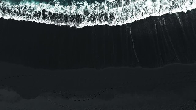 Aerial footage of white ocean waves crashing and receding on a dramatic black volcanic sand beach, moody cinematic atmosphere