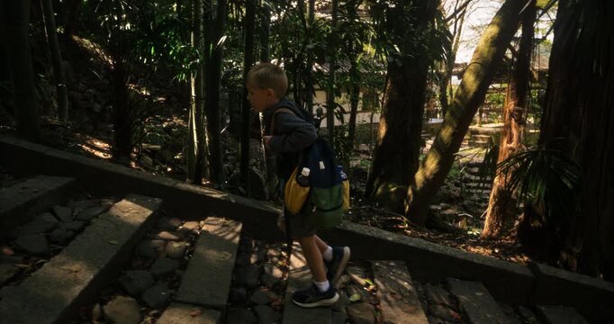 Little boy with backpack walking up stone steps in forest near old Japanese village - tracking shot