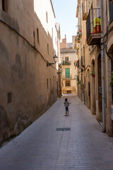 Obraz premium Back view of a small child walking down a quiet, sunlit cobblestone street lined with tall ancient buildings and traditional balconies in a historic Mediterranean town.