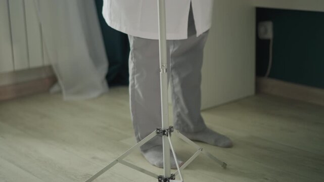 Home nurse setup. Adjusting intravenous stand at home. Medical professional prepares infusion bedside at residence. Nurse arranges IV equipment at home with careful attention to detail
