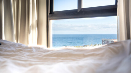 Fototapeta premium First person perspective from a bright white bed looking through a window at the beautiful blue ocean horizon.