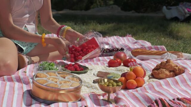 Woman preparing fresh raspberries for a summer picnic on a striped blanket in a park with fruits and pastries