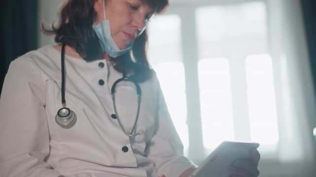 Caucasian physician using tablet near window light, professional and calm. Stethoscope draped over coat, mask lowered, fingers swipe screen while reviewing charts, soft sunlight through curtains,