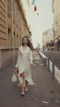 Beautiful woman wearing a chic white lace dress and holding a straw hat walking confidently along a charming, sunlit european street, her long hair flowing as she moves toward the camera, slow motion