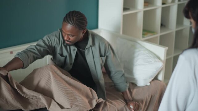 Black man seated on bed avoiding nurse, pulling blanket up and shielding face, female nurse standing by bedside in soft clinical coat, domestic shelving background, tense but caring homecare