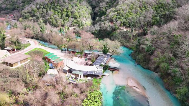 Aerial tracking shot moving forward over the vibrant turquoise river curves, finishing with a smooth pan down directly onto the crystal-clear pool of the Blue Eye water spring in Albania