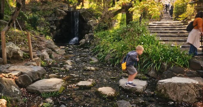 Boy with backpack hopping over stone steps of stream in old Japanese village - steady cam shot forward
