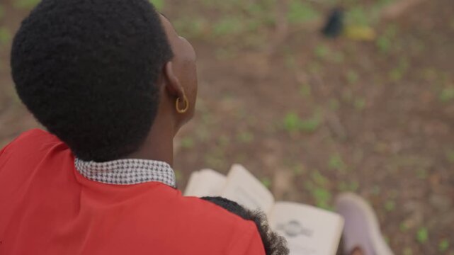 Black mother daughter reading outdoors intimate back view of mother in red jacket with child on lap, open book on knees, soft daylight, grassy park ground, gentle nurturing mood, quiet connection