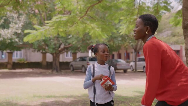 Playful park sendoff mother and daughter sharing snack under tree, mother leans in to kiss daughters cheek while handing lunchbox, lighthearted morning routine, warm smiles, casual clothing, suburban