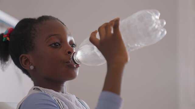 Black daughter drinking water from bottle with focused expression, clear plastic bottle and casual home setting, postplay hydration and healthy habit, closeup hand to mouth, refreshing sip