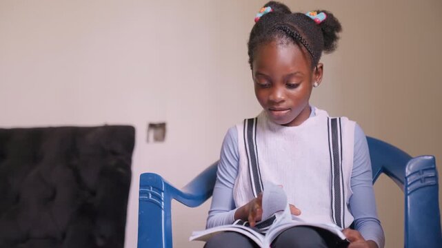 Smiling African girl relaxes with favorite book. Cheerful young girl immersed in intriguing story on soft chair. Joyful African American girl comfortably reading engaging story with bright smile
