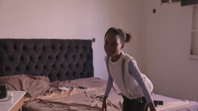 Childly girl moves freely over bedding. Cheerful young girl twirling amidst rumpled sheets. Ebullient black daughter joyfully dancing in casual attire amidst messy sheets with warm sunlight