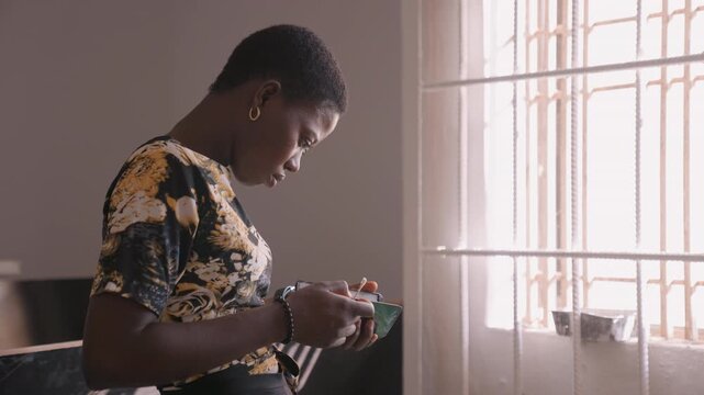Black woman checking phone by window, soft morning light filters through blinds, focused expression while inspecting device, hoop earring glinting, hands adjusting strap, quiet domestic interior,