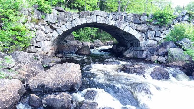 Drone moving upstream through a low and narrow old stone bridge surrounded by a fresh green forest on late sunny spring afternoon.