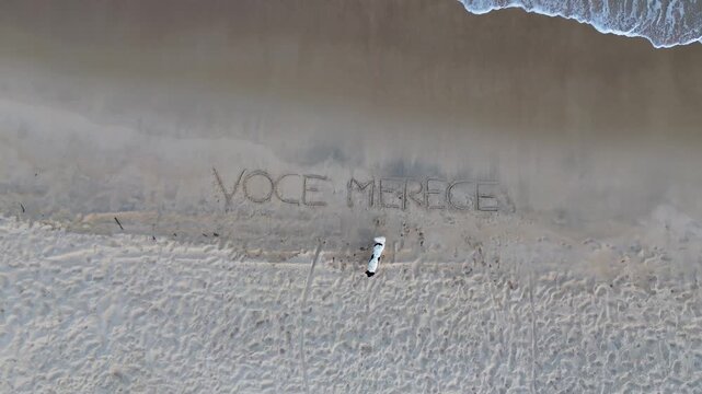 The phrase "voce merece" marked in the sand of a beach