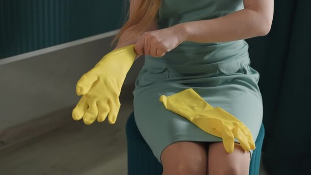Homeowner putting on yellow gloves, sitting on stool in cozy interior, soft natural light, green dress, hands fastening rubber fit, slow deliberate movements, morning cleaning prep, tidy domestic