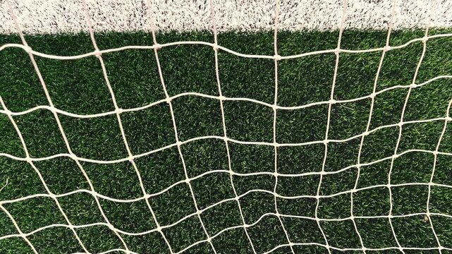 Tight, stationary close-up looking through the grid of a white soccer goal net as it blows dynamically in strong wind against a beautifully blurred green turf background.