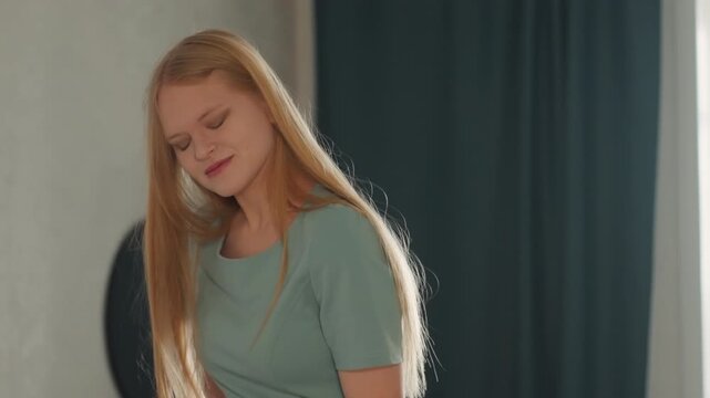 White woman house cleaning, satisfied smile and relaxed posture, sunlight on long blonde hair, teal top and curtain background, tidy livingroom and sense of completion, calm confident expression