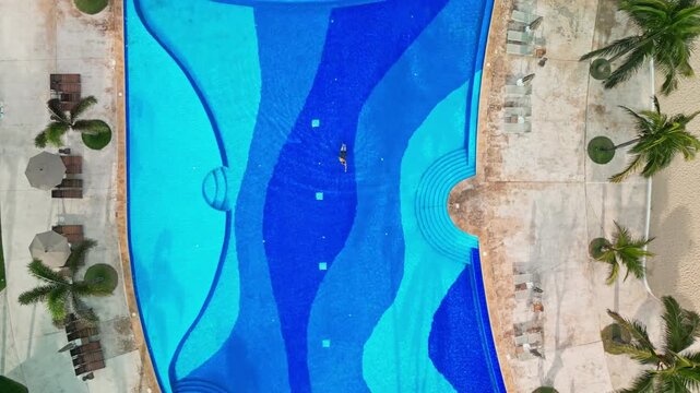 Aerial overhead pull back over Nuevo Vallarta resort pool with wave pattern and lone swimmer. Horizontal video.