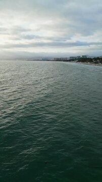 Aerial drone dolly approaching Nuevo Vallarta resort coastline from Pacific Ocean. Rough sea, palapas, hotel, mountains. Cloudy to clearing sky. Vertical video.