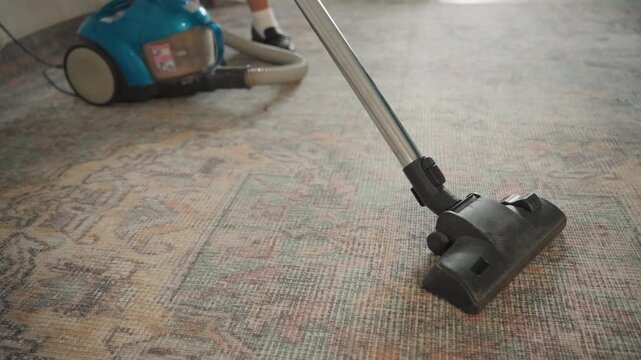 Closeup vacuum cleaning patterned carpet, blue canister vacuum gliding over textured rug, gentle suction lifting dust and fibers, soft morning light, quiet domestic chore capturing satisfying motion