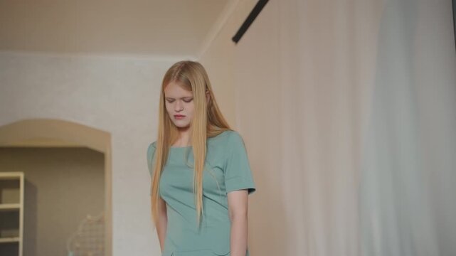 White woman cleaning house, standing pensively in sunlit living room blonde hair and teal dress, curtain and shelving in background, subtle mess from chores, homeowner pausing between tasks, domestic