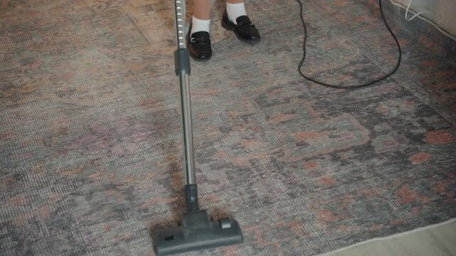 Closeup vacuuming rug in house showing homeowner cleaning patterned carpet, cord trailing across floor, black shoes with white socks visible, steady backandforth motion lifting dust from textured