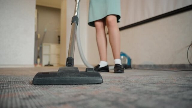 Lowangle vacuum cleaning house carpet young helper legs in skirt and socks, canister vacuum head gliding over patterned carpet, hallway interior, morning sunlight casting soft shadows, methodical