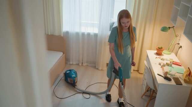 Girl vacuuming bedroom in house, student tidies desk, plugs vacuum, moves canister across wood floor, sunlight through curtains, cozy decor with lamp and plant, playful yet focused mood, morning
