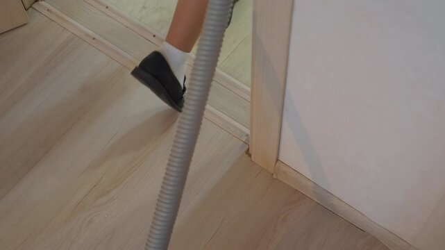 Woman vacuuming house hallway floor, closeup of lower legs in teal skirt and black shoes, compact canister vacuum with hose navigating doorway threshold on wooden laminate surface, domestic cleaning