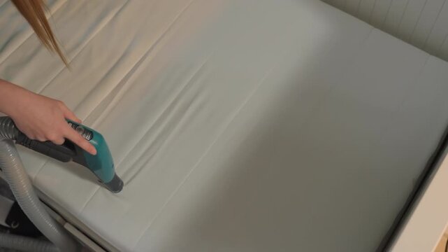 Caucasian woman cleaning mattress surface using handheld extractor vacuum, homeowner carefully removes stains and dust from fabric with nozzle glide, closeup of motion on bed in quiet bedroom,