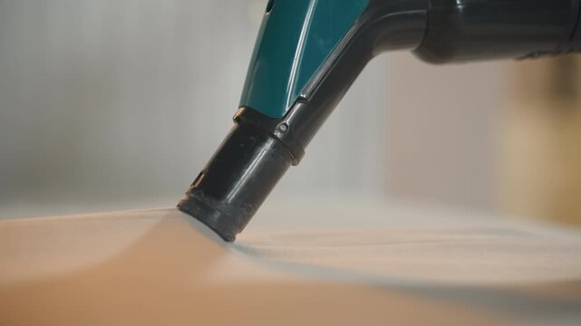 Close up mattress cleaning vacuum nozzle, teal handheld upholstery attachment gliding over fabric surface, gentle suction lifting dust and debris from mattress in warm bedroom interior, soft shallow