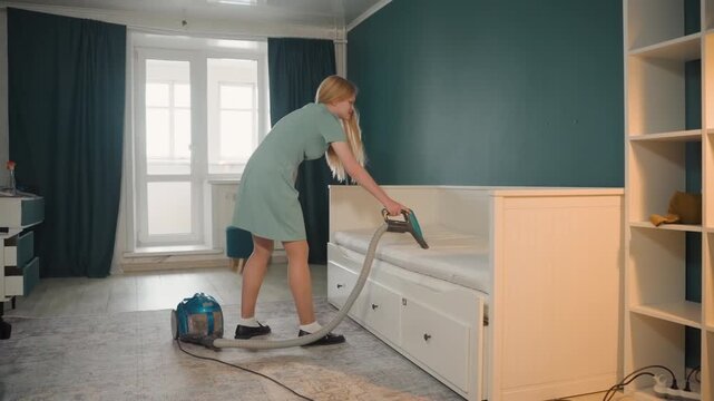 White woman cleaning bedroom with vacuum inside house. Homemaker vacuuming daybed and mattress seams, pulling dust from upholstery, moving vacuum canister across carpet, adjusting curtains,