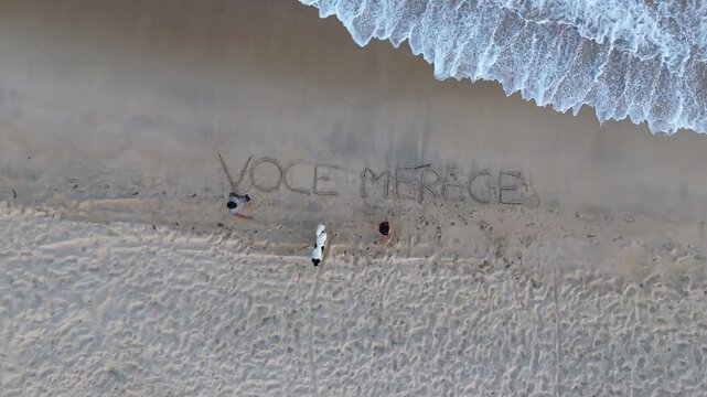 A young couple and a dog on a Brazilian beach with an inscription marked in the sand that says "voce merece" (you deserve).