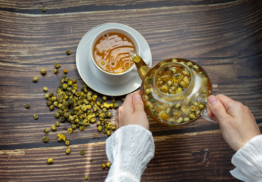 Top view of female hands pouring freshly brewed chamomile and chrysanthemum tea from a glass teapot into a cup. Wellness, relaxation and healthy lifestyle concept.