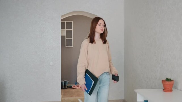 Caucasian woman enters minimalist home study, carries notebook and laptop, sets items on desk, adjusts lamp and plant, prepares online grammar tutoring session with calm focused expression