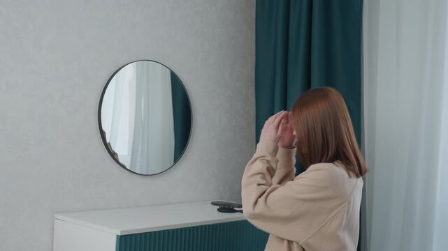 Caucasian student checking mirror reflection in bedroom with teal curtains, studies posture and expression before online lesson, adjusts hair and sweater, introspective soft light mood