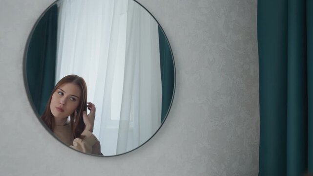 Young woman preparing in mirror, checking hair and makeup by window light, quiet bedroom scene with round wall mirror and teal curtain, hesitant expressions shift to deliberate grooming as she brushes