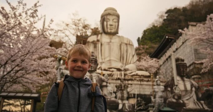 Smiling blond boy in front of ancient, large, stone buddha statue surrounded by cherry blossoms