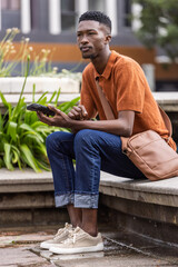 Obraz premium Adult man sitting on concrete ledge in urban plaza holding smartphone and wearing rust-orange polo
