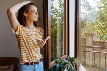 Obraz premium A woman in striped shirt stands by an open window, basking in daylight, lost in thought. A cozy home scene for lifestyle, relaxation, and simple living.