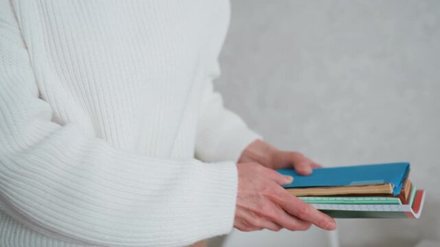 White woman stacking books on desk, wearing cozy sweater beside succulent pot, hands arranging notebooks and journals on tidy workspace, focused preparation for lesson or study routine with minimal