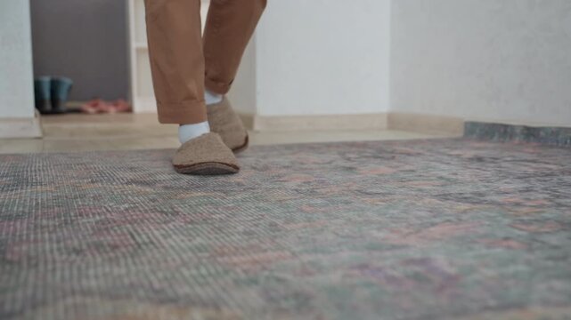 Slippers on patterned rug near doorway. Casual tutor walks into cozy home study wearing beige pants and white socks, pauses by desk, soft daylight highlights vintage rug texture and calm morning