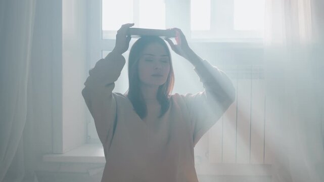 Young woman meditating near window, balancing book on head while practicing posture and silent grammar drills, soft sunlight rays across cozy living room, crosslegged on rug in sweater and jeans, calm